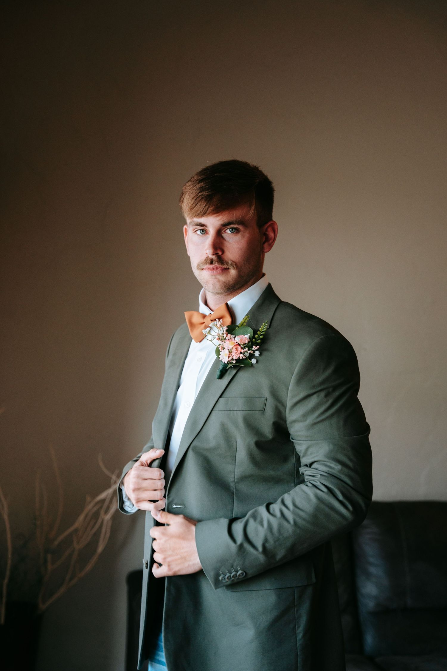 Dapper groom in olive suit with orange bow tie and floral boutonniere posing indoors.