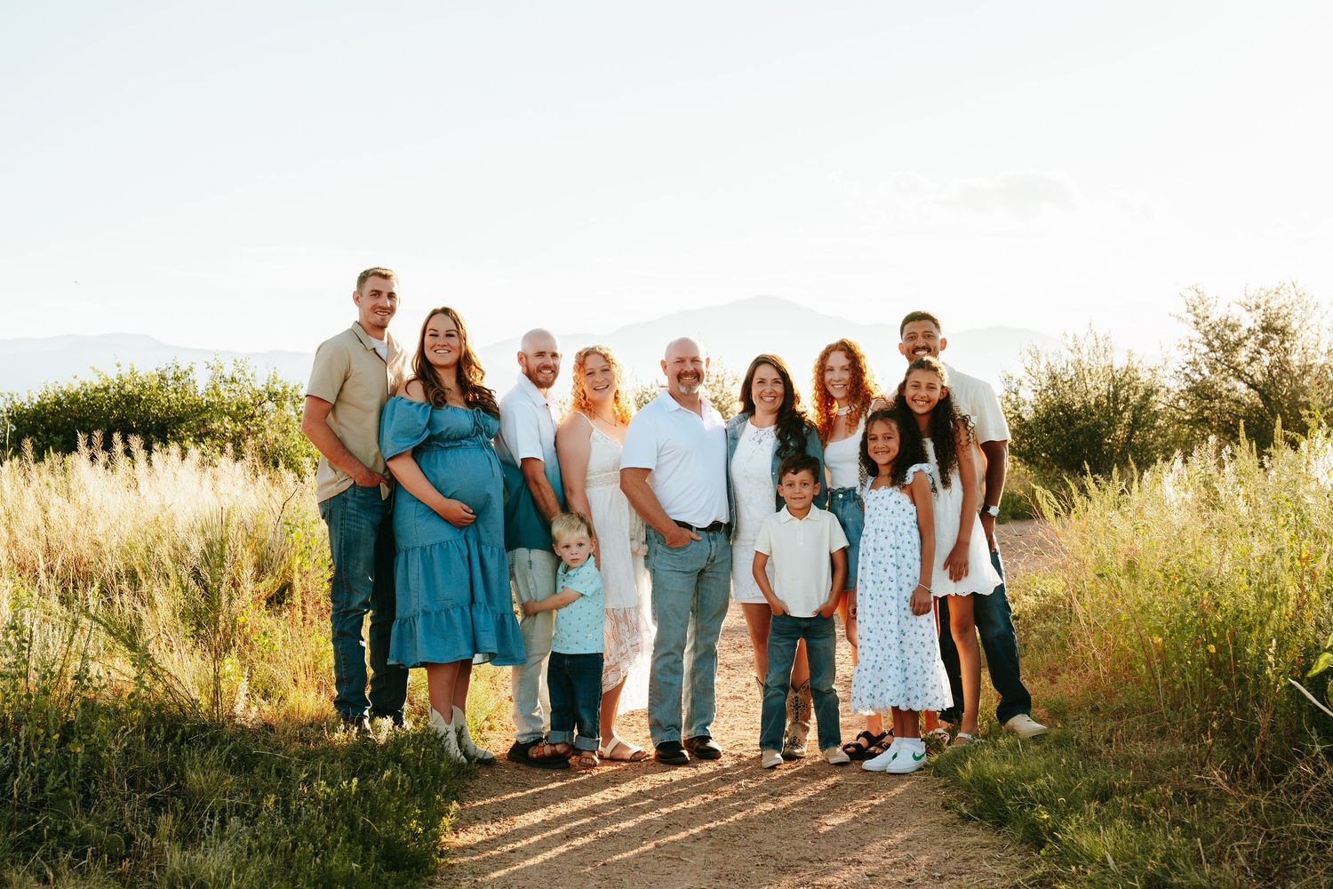 Large extended family group portrait outdoors on sunny day, posing together on dirt path with green fields behind.