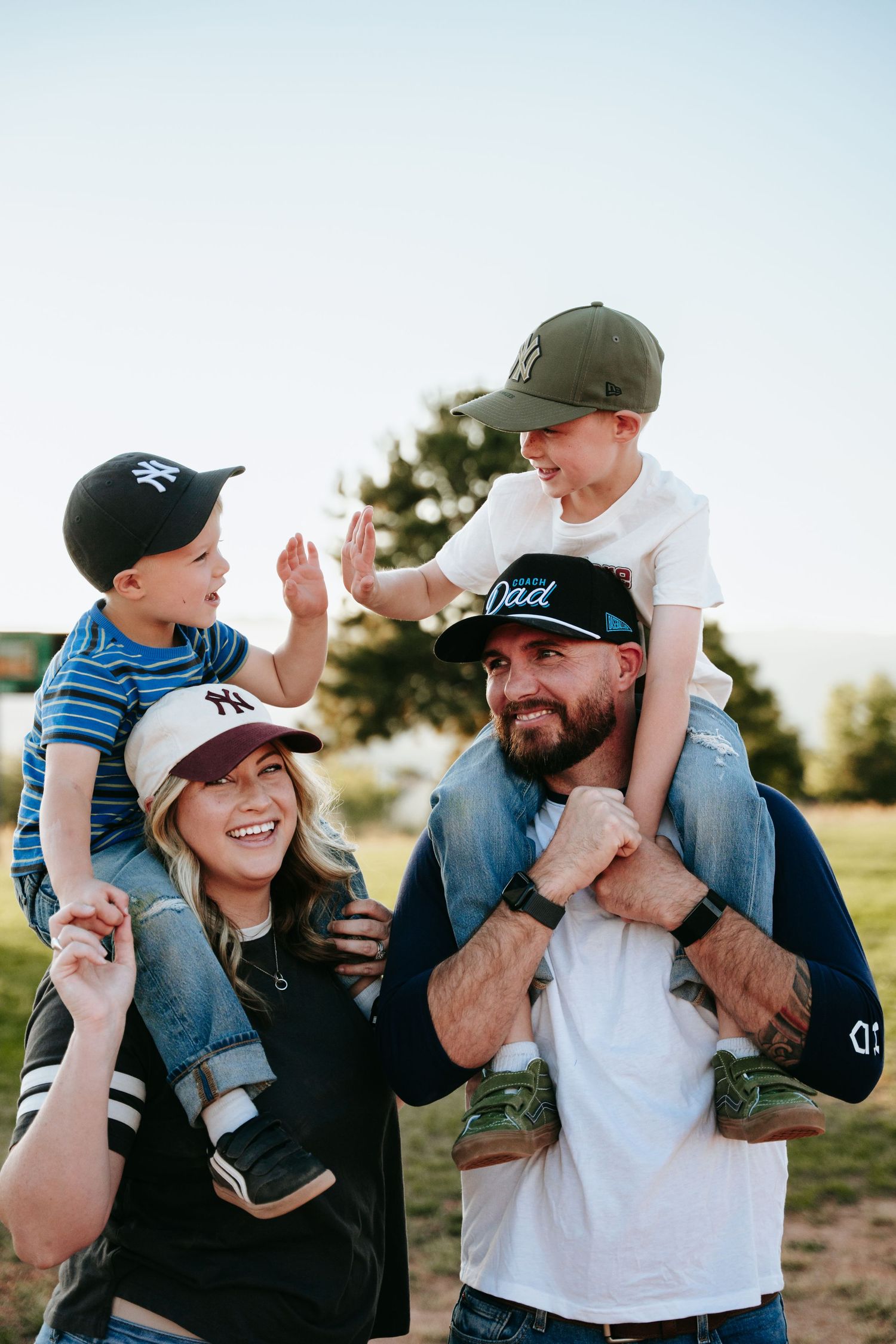 Smiling couple carrying two laughing young boys on their shoulders outdoors, all wearing baseball caps, in a sunny grassy park.
