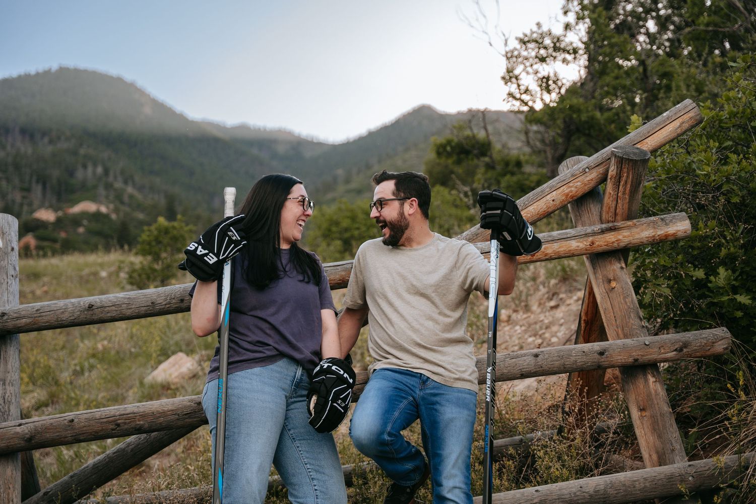 A smiling couple in work gloves leaning on a rustic wooden fence with mountains and trees in the background.