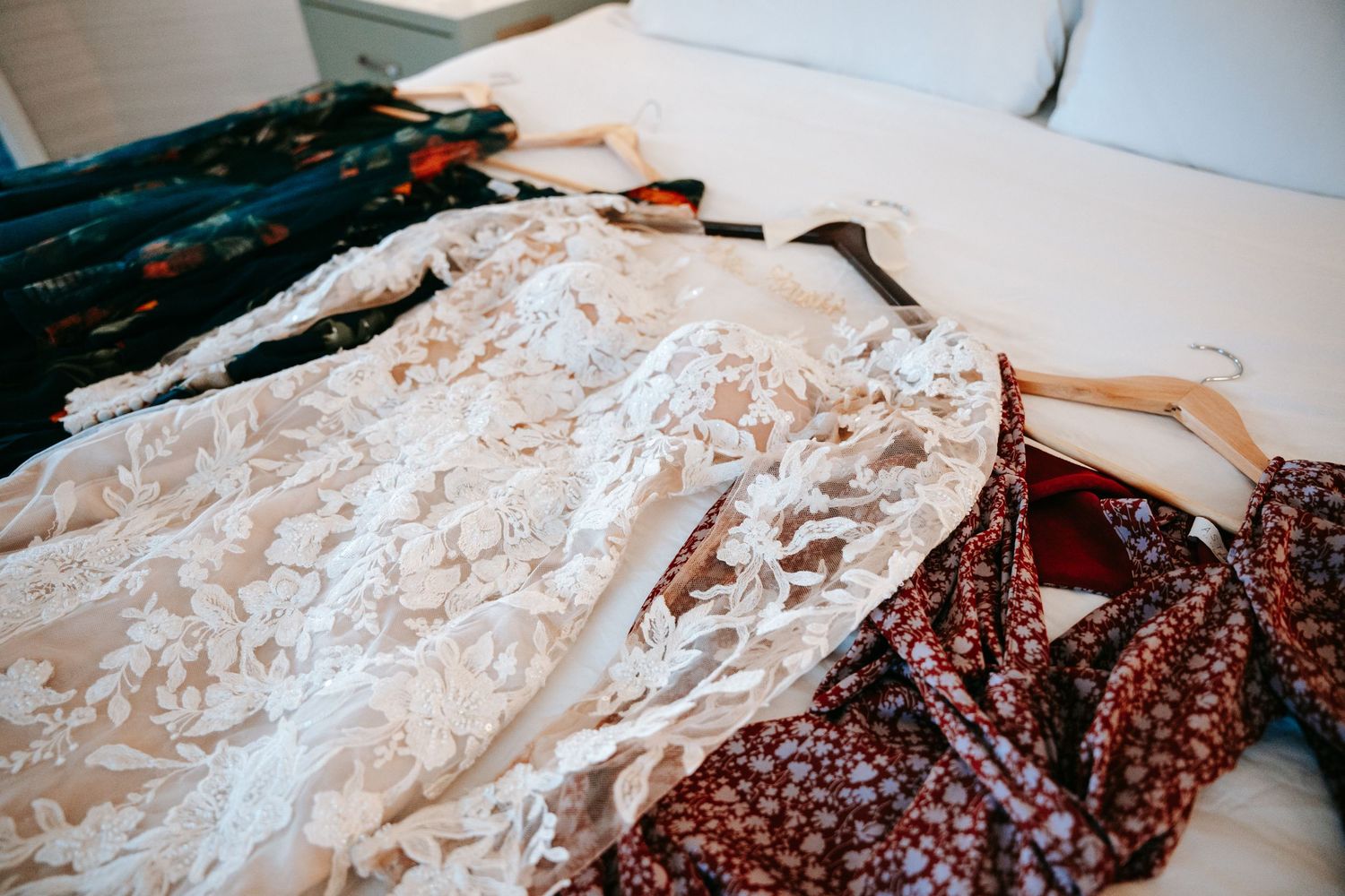 Elegant lace wedding gown and floral bridesmaid dresses displayed on hangers on a white bed.