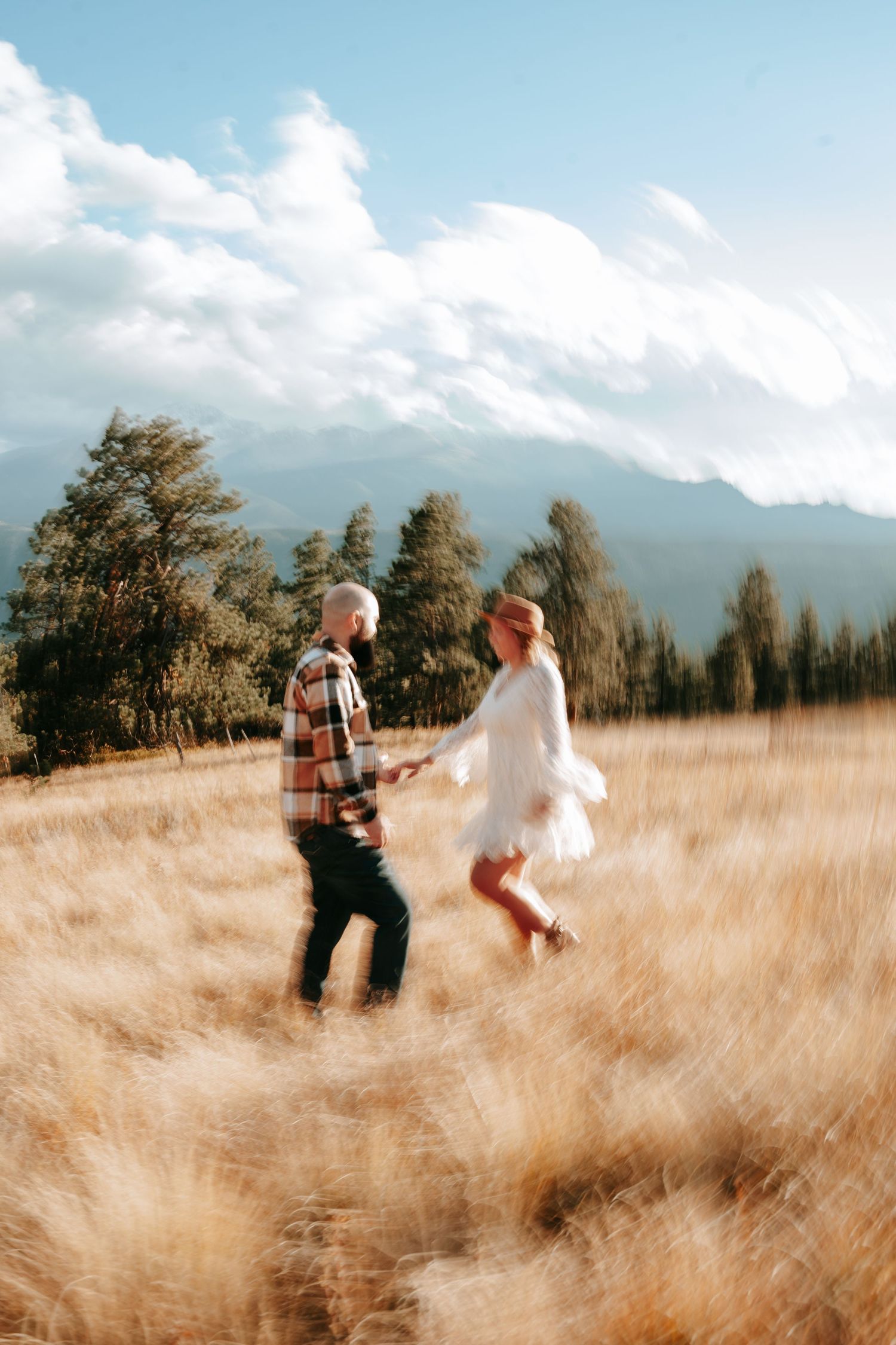 Bride in white dress and groom in plaid shirt walk through golden meadow with mountain backdrop in motion-blur photo.