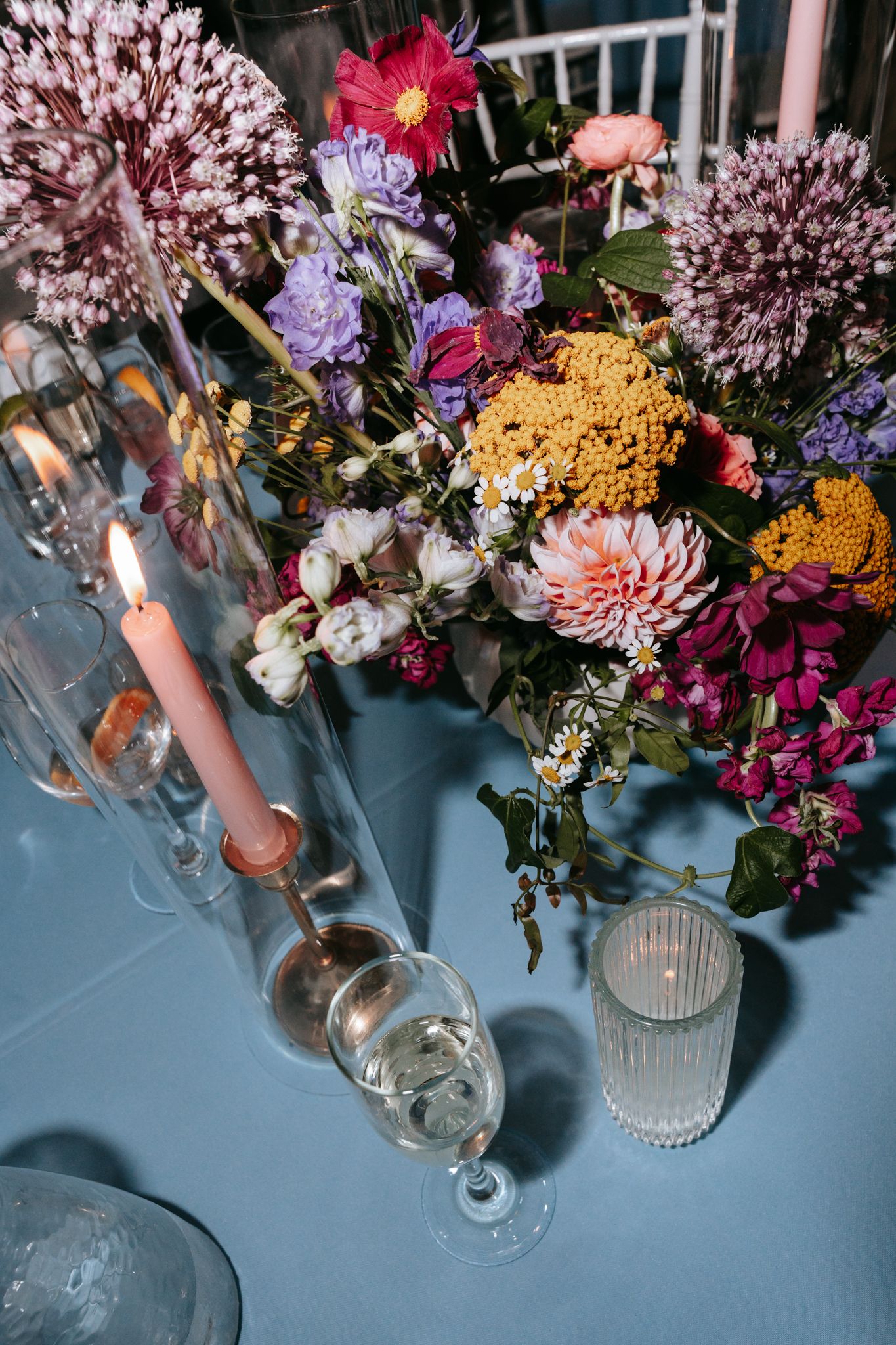 Colorful wedding table centerpiece with vibrant mixed flowers, candles, and glassware on blue linen.