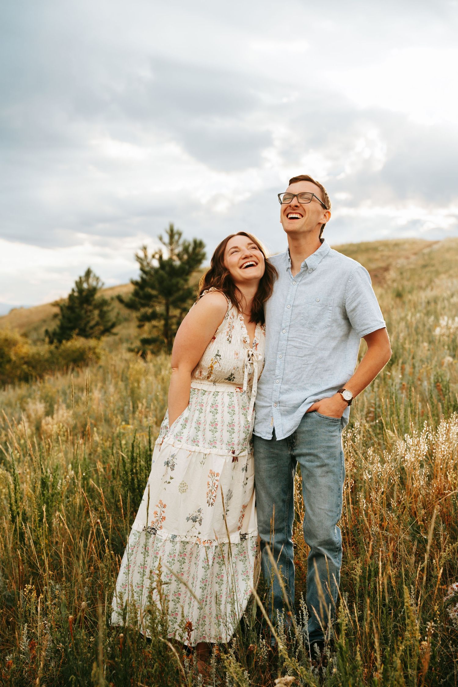 Happy couple laughing and looking up together while standing in a golden meadow under cloudy skies.