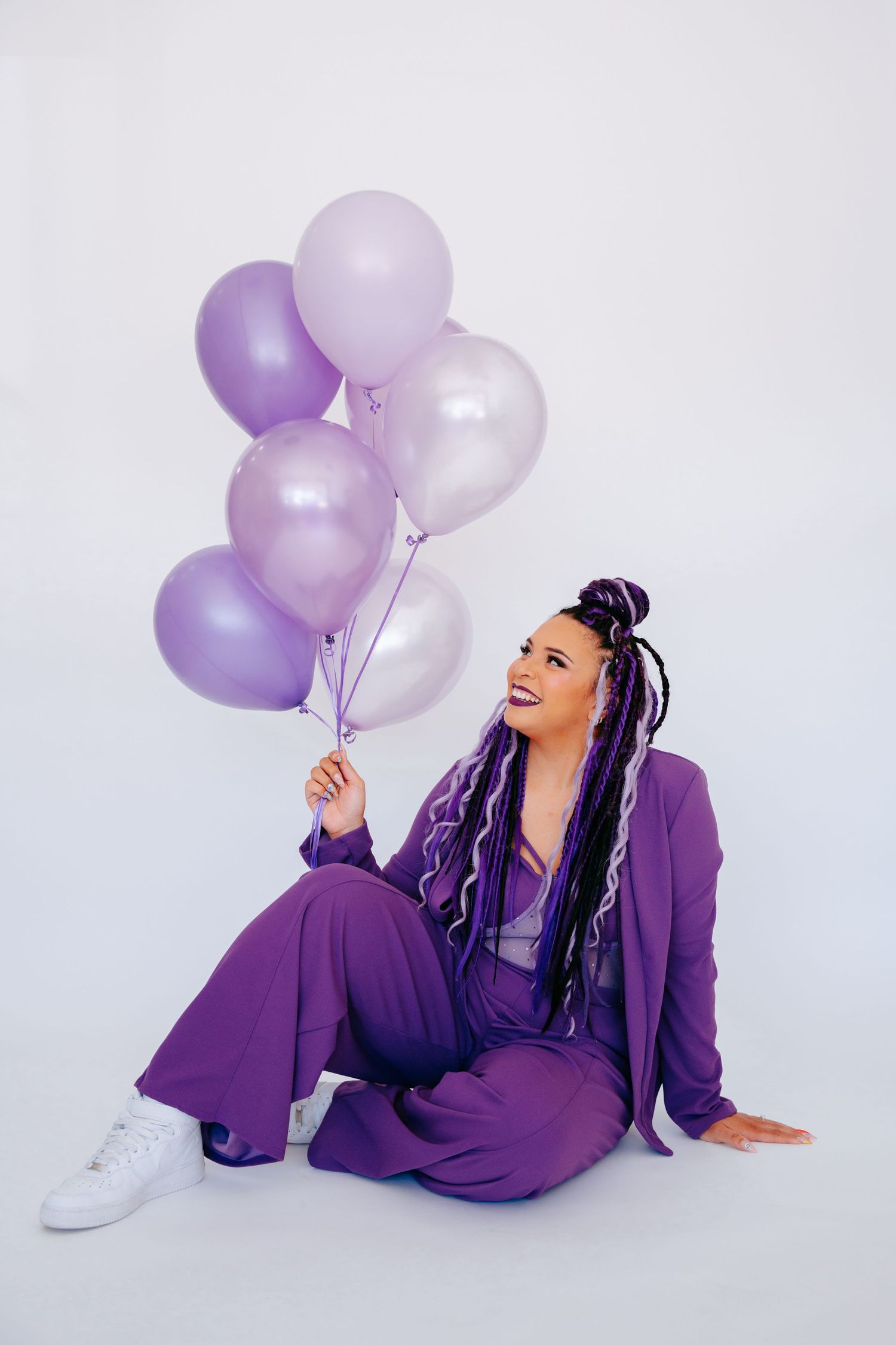 Smiling woman in purple suit sitting on floor holding bunch of lavender and pearl balloons on white background.