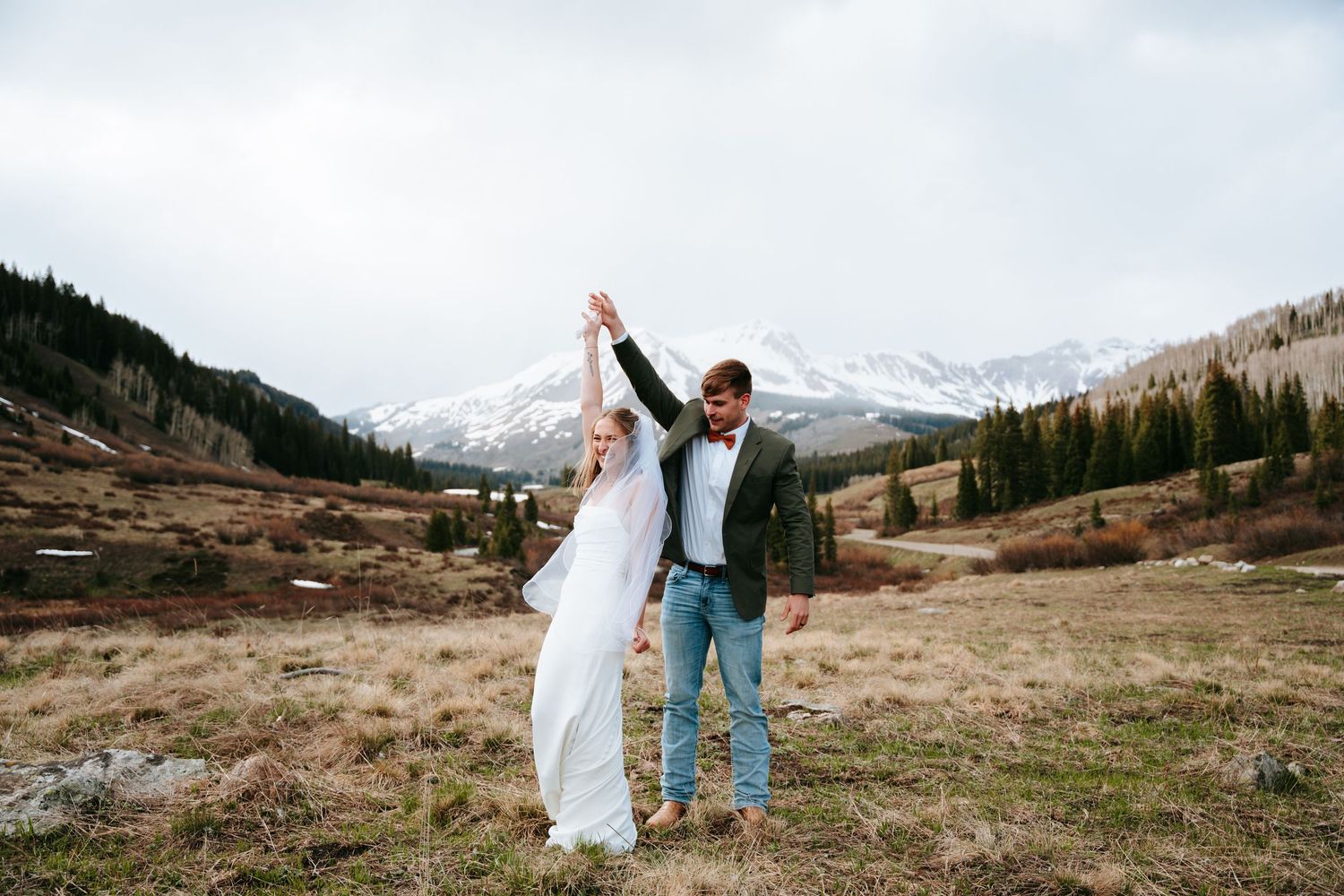 Groom twirls bride in a mountain meadow with golden trees and snow-covered peaks in the background.