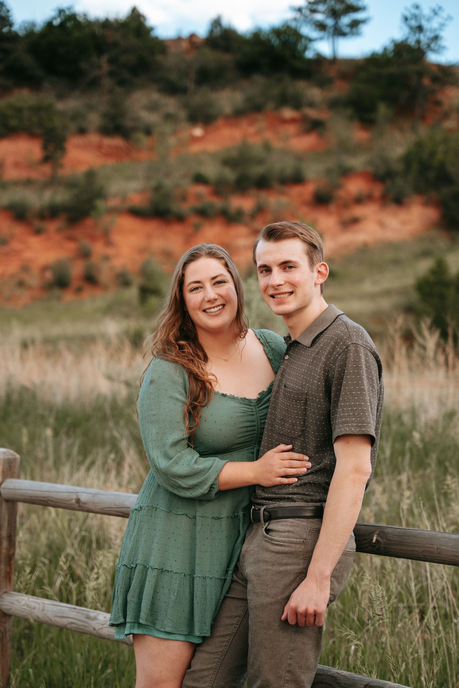 A smiling couple poses together near a wooden fence with red clay hills and green meadows in the background.