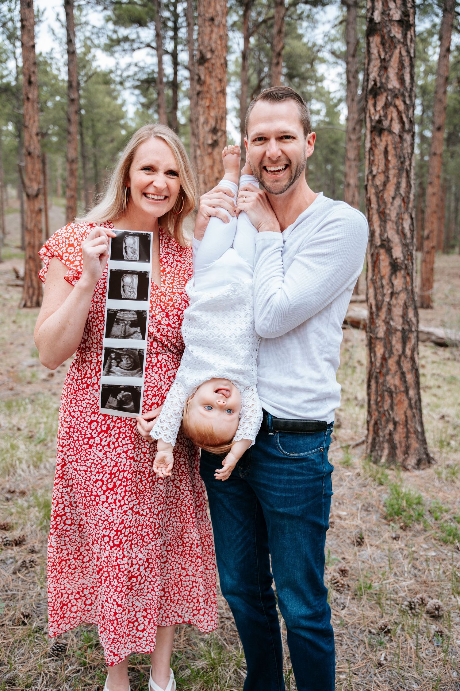 Happy couple holding ultrasound photos announces pregnancy while man holds toddler upside down in pine forest.