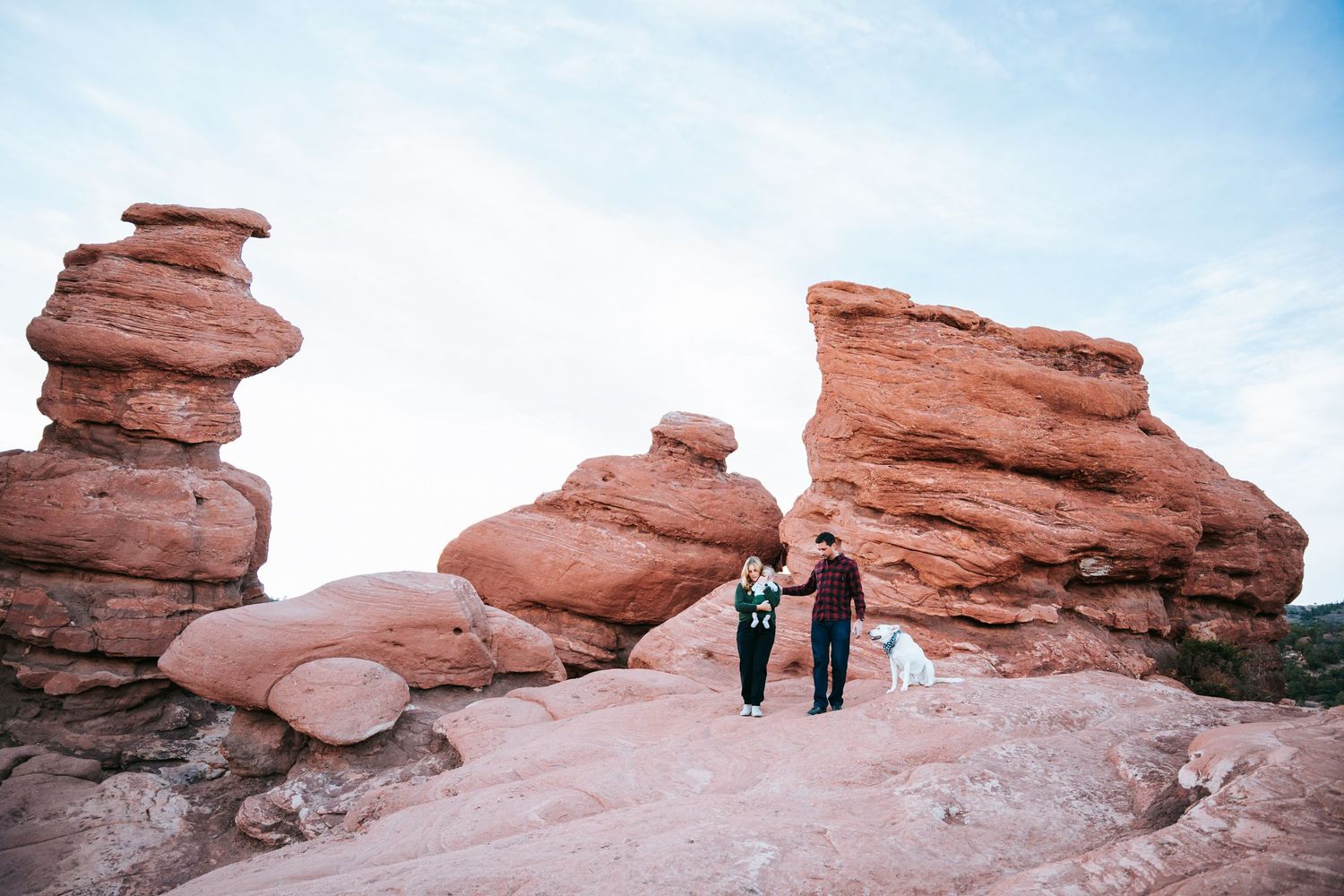 Couple with white dog standing between towering red rock formations at Garden of the Gods.