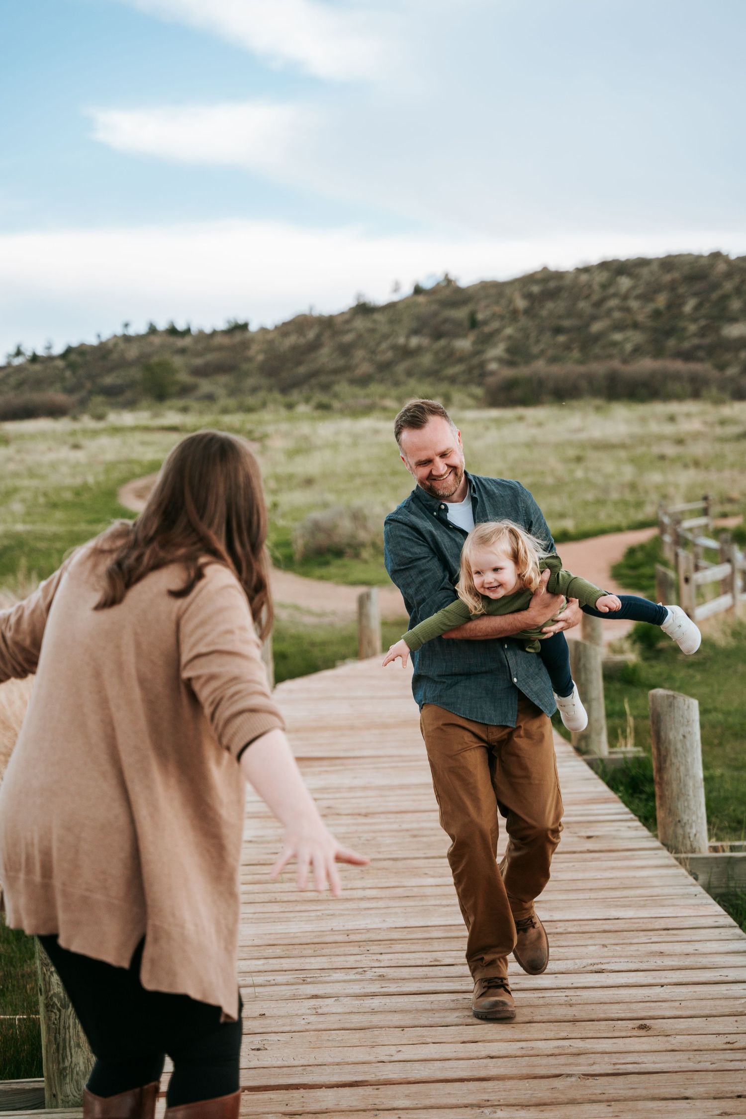 Happy father carrying laughing child on a wooden boardwalk path through green meadow toward mother.
