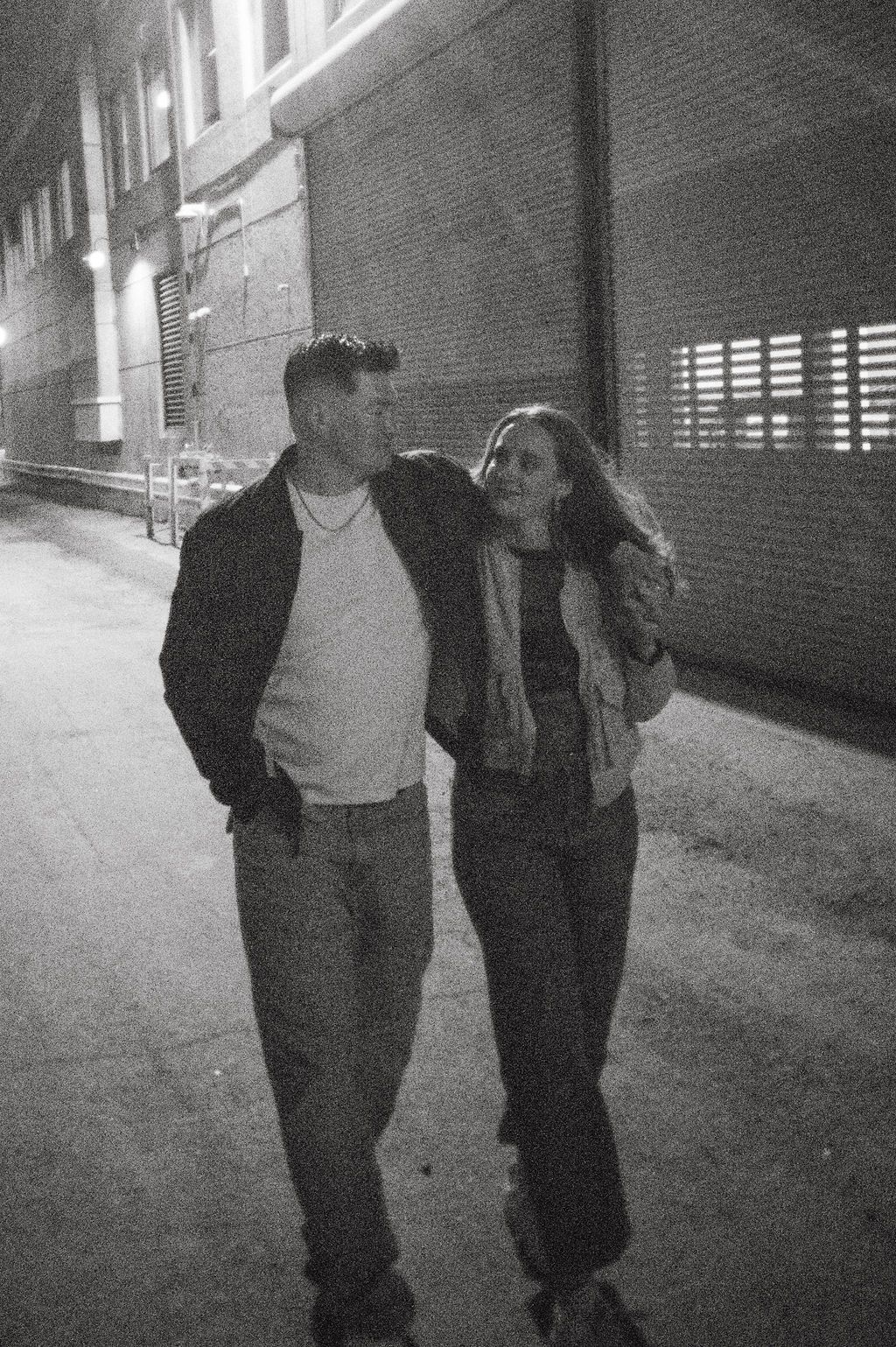Black and white photo of couple walking together at night along urban brick alleyway.