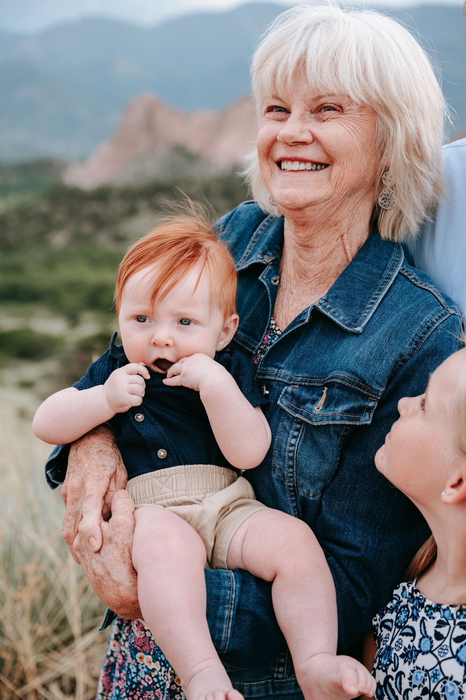 Smiling grandmother holding red-haired baby with young girl beside her near rocky mountains.