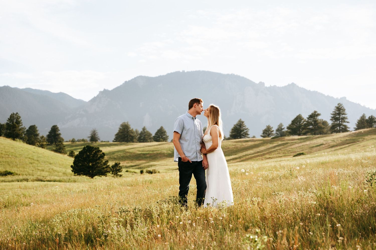 Couple sharing a kiss in sunlit mountain meadow with rocky peaks and pine trees behind them.