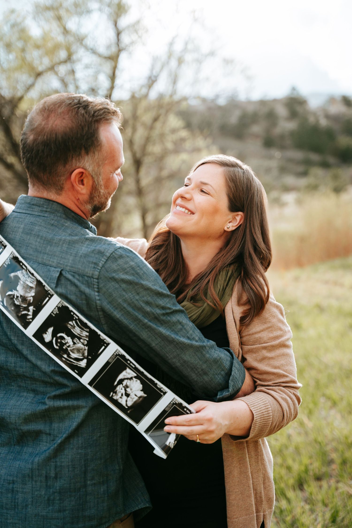 Smiling woman hugging man holding ultrasound photos during outdoor pregnancy announcement session.