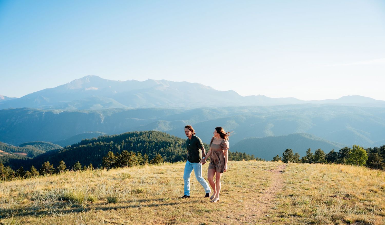 Couple walking hand in hand through a golden meadow with expansive mountain range behind them.