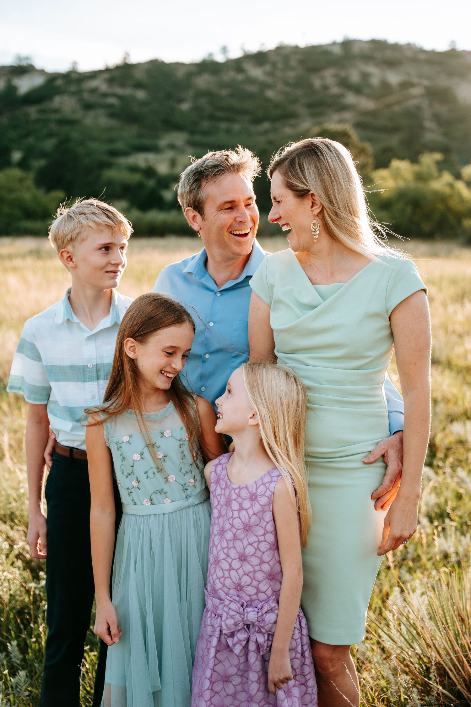 Joyful family of five in coordinating mint and blue outfits laughing in a golden meadow.