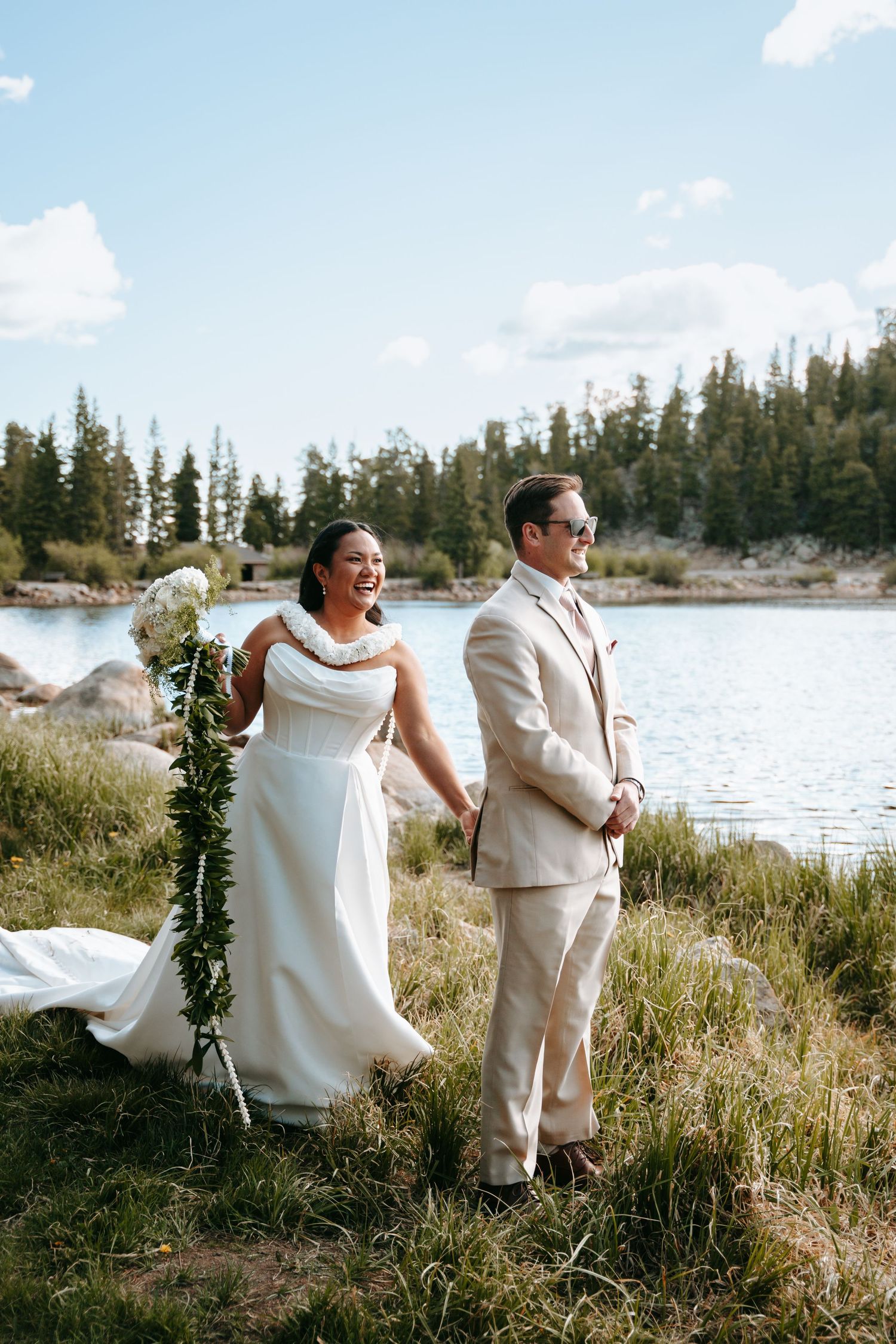 Bride in white gown and groom in beige suit hold hands by a mountain lake surrounded by pine trees on their wedding day.