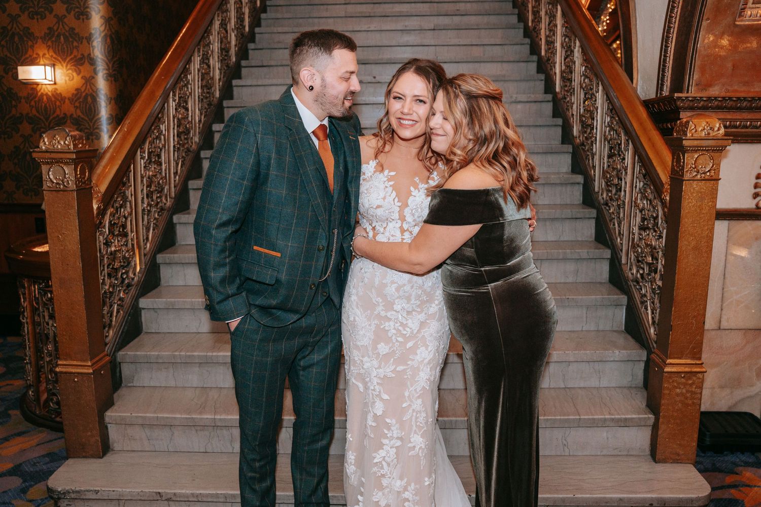 Bride in lace gown embraces groom and guest on elegant wooden staircase.
