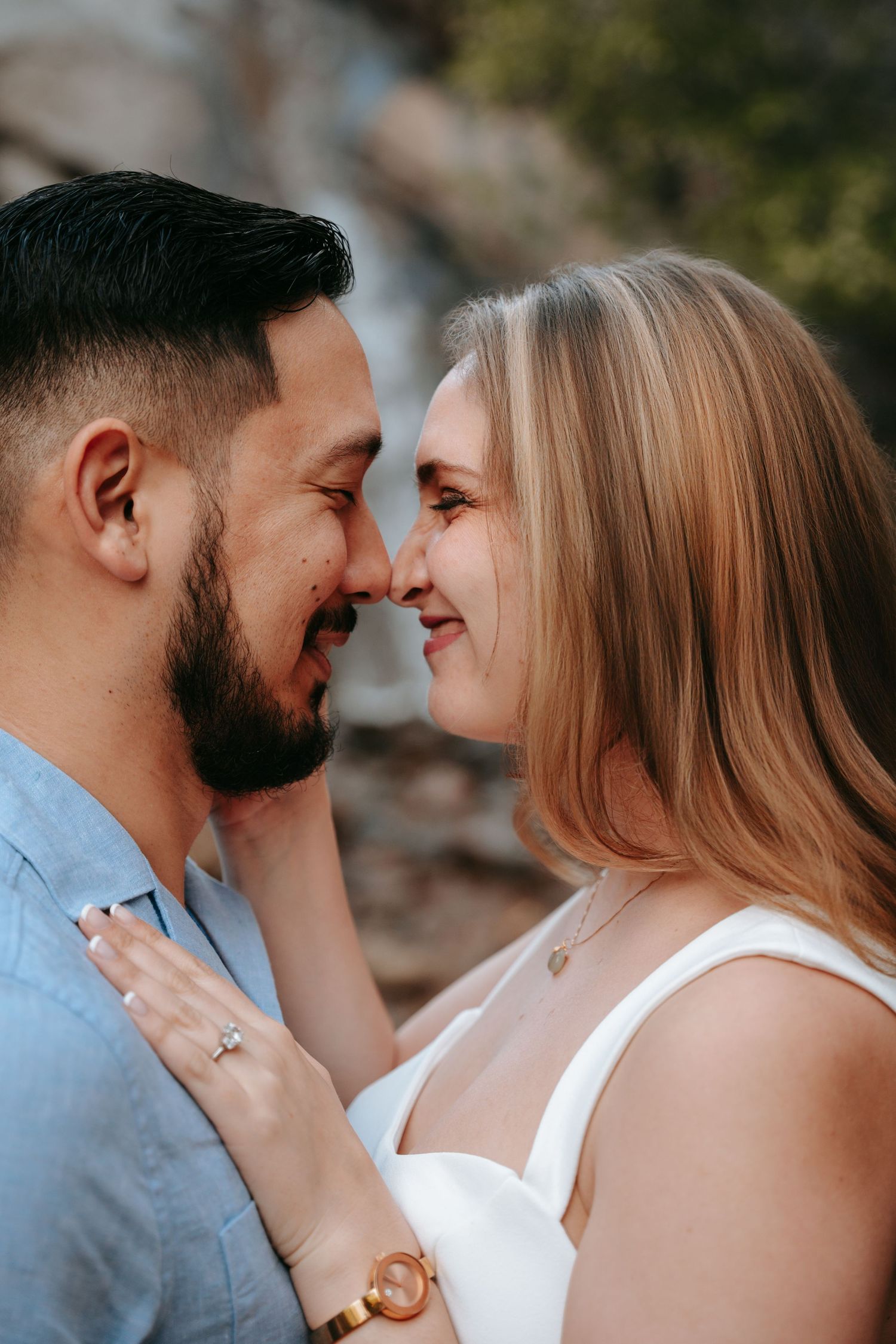 A couple shares a romantic nose-to-nose moment, the woman wearing an engagement ring and white dress, the man in a blue shirt.