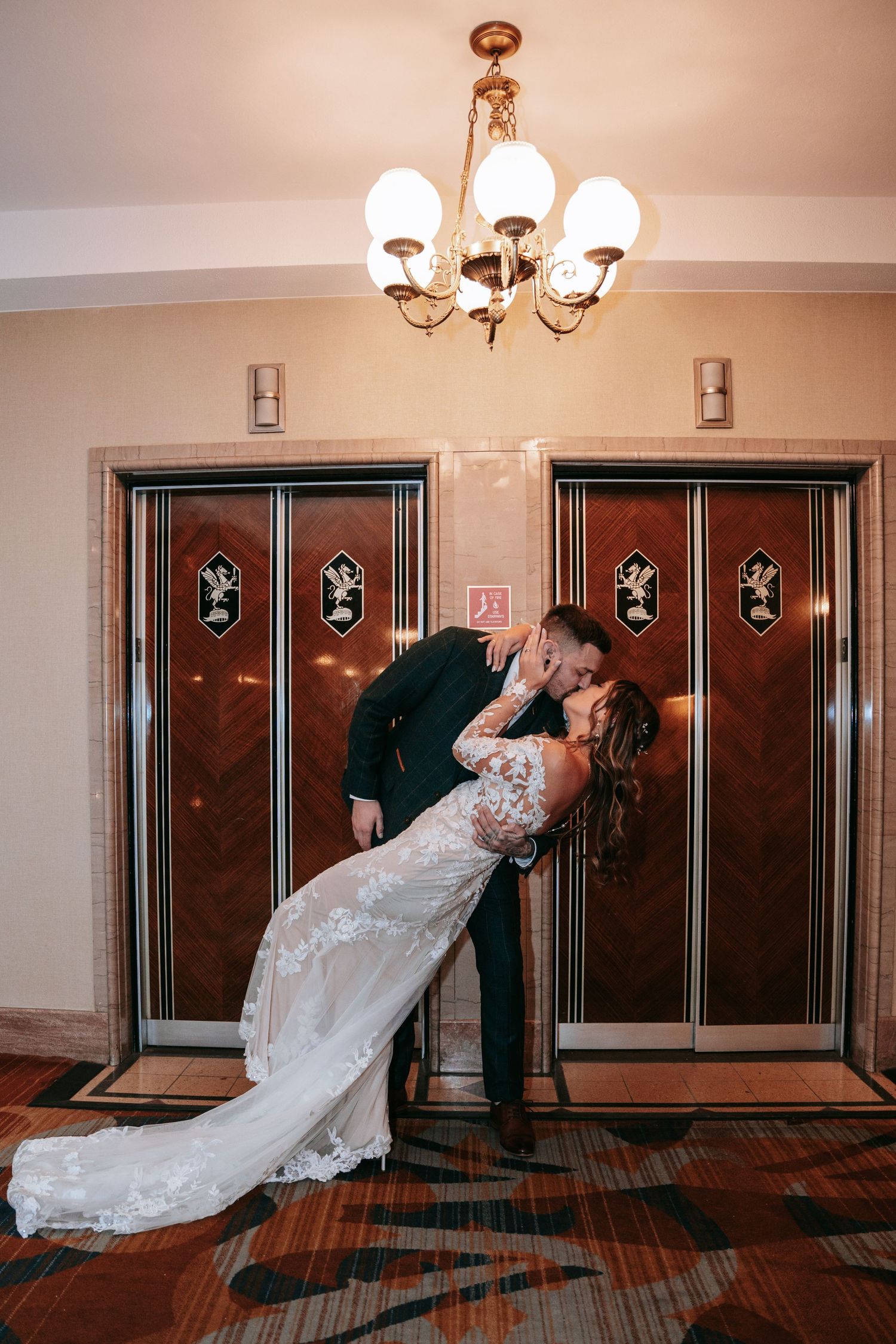Bride and groom sharing a romantic dip kiss in front of art deco elevator doors under chandelier.