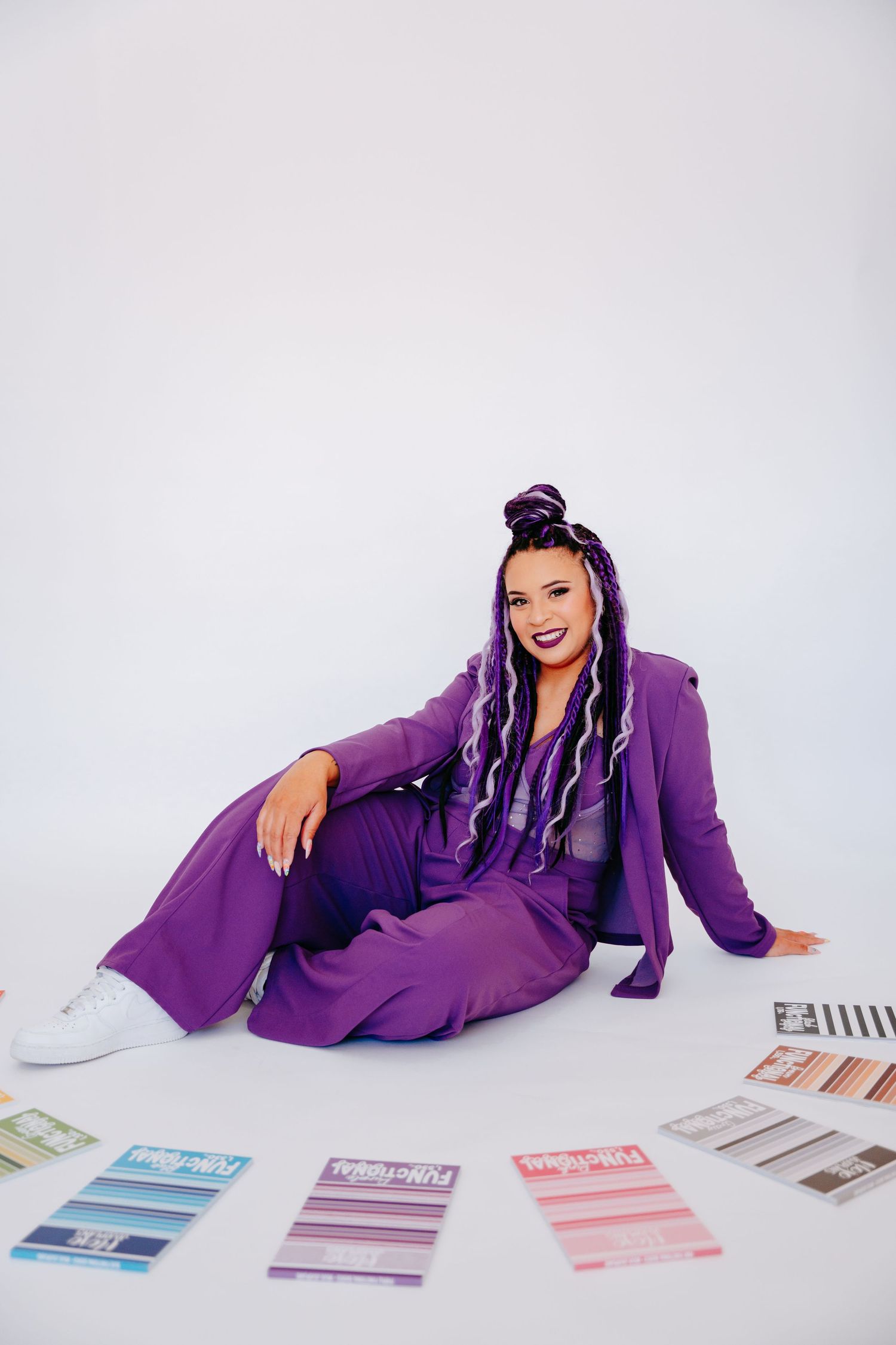 Woman in purple suit with long braids sitting on white floor smiling, surrounded by colorful design swatches.