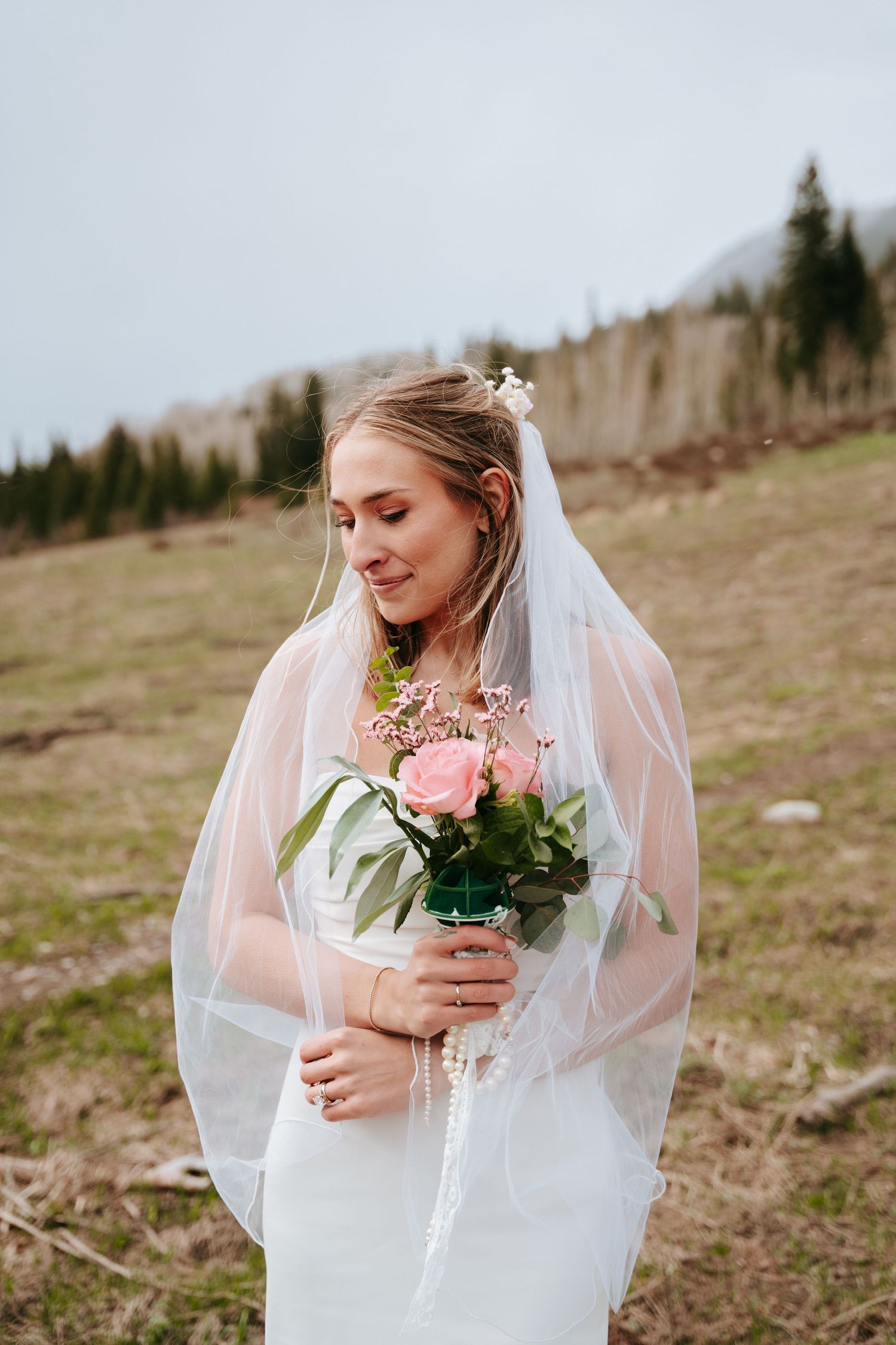 Beautiful bride in white gown holds pink rose bouquet while her veil drapes softly in the breeze.