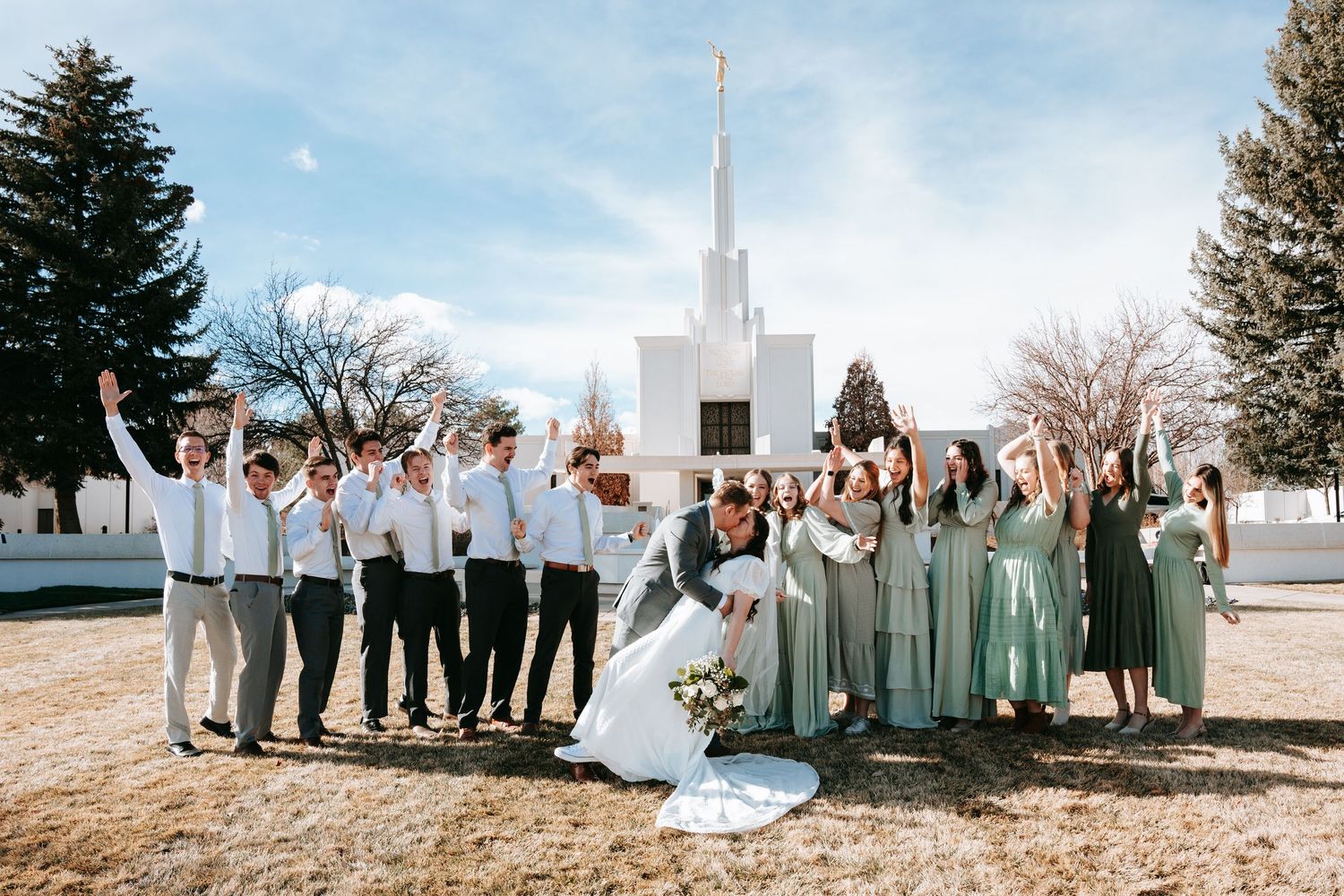 Wedding party celebrates outside a white LDS temple, bridesmaids in sage green dresses with groomsmen in gray suits.