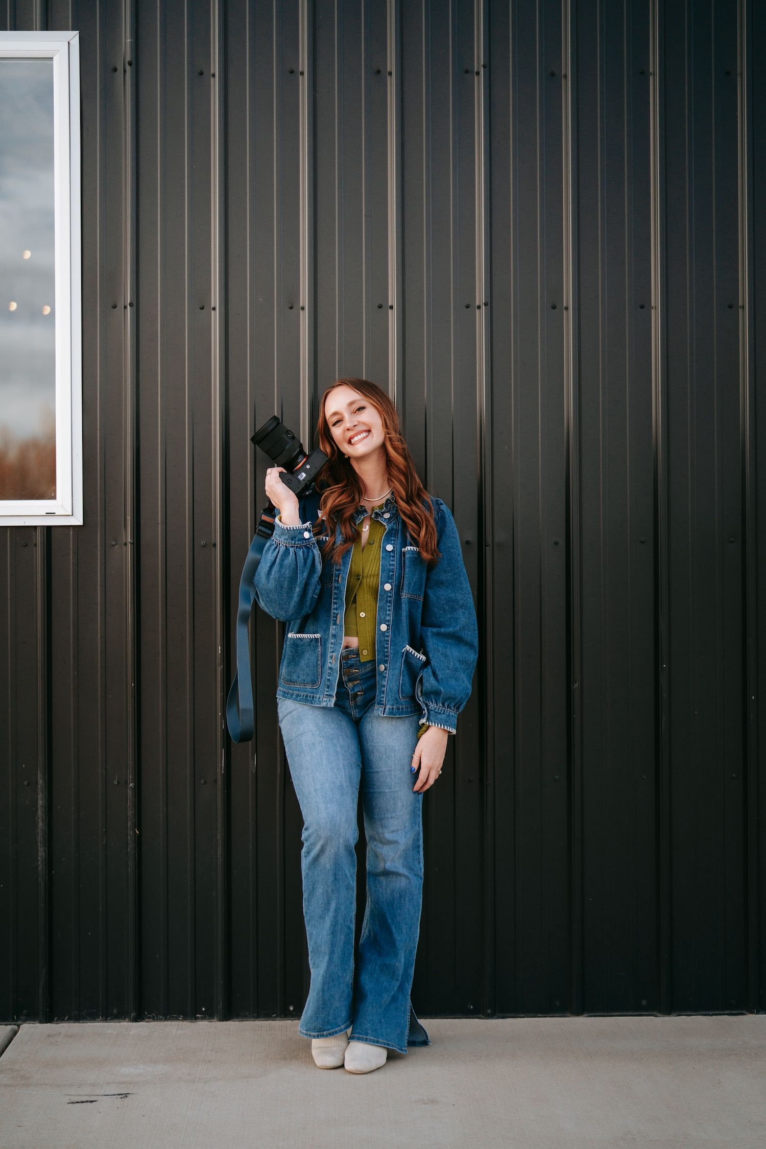 Smiling redhead photographer in denim jacket holding camera against dark metal wall.