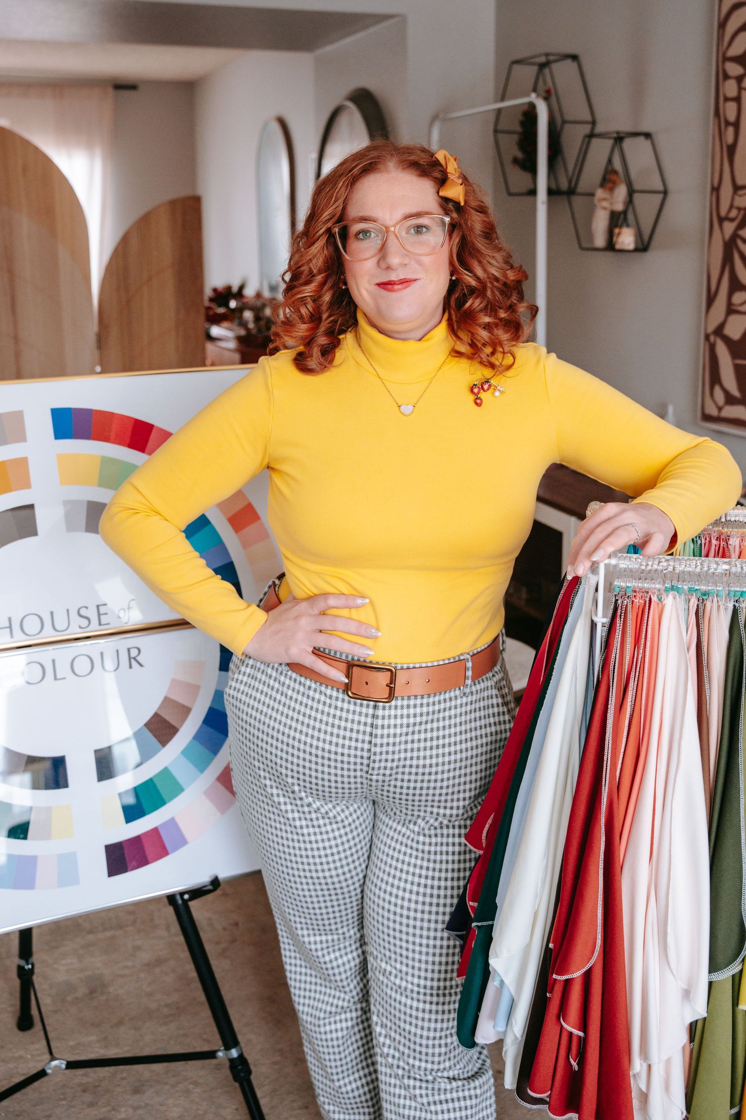 Redhead woman in yellow turtleneck posing beside House of Colour wheel and fabric swatches.