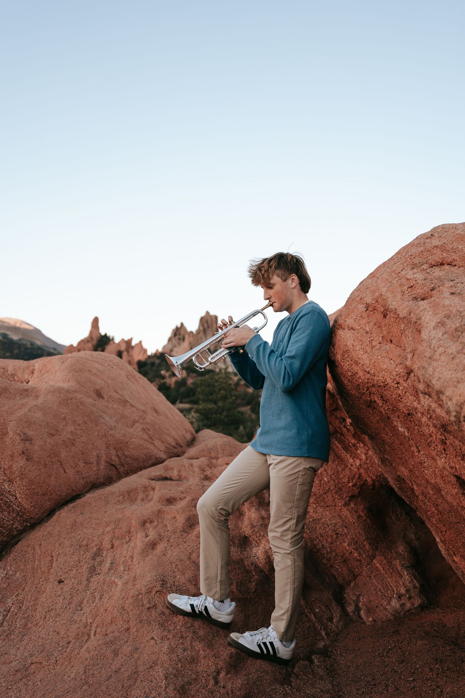 Young man playing silver trumpet leaning against red sandstone rocks at Garden of the Gods.
