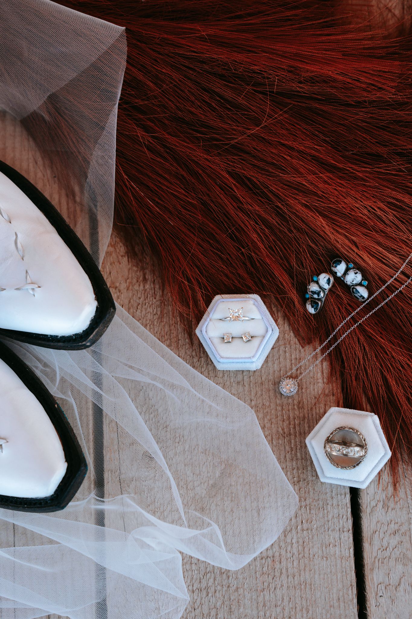 Flat lay of wedding rings in white hexagon boxes, veil, black heels, and red hair on wooden surface.