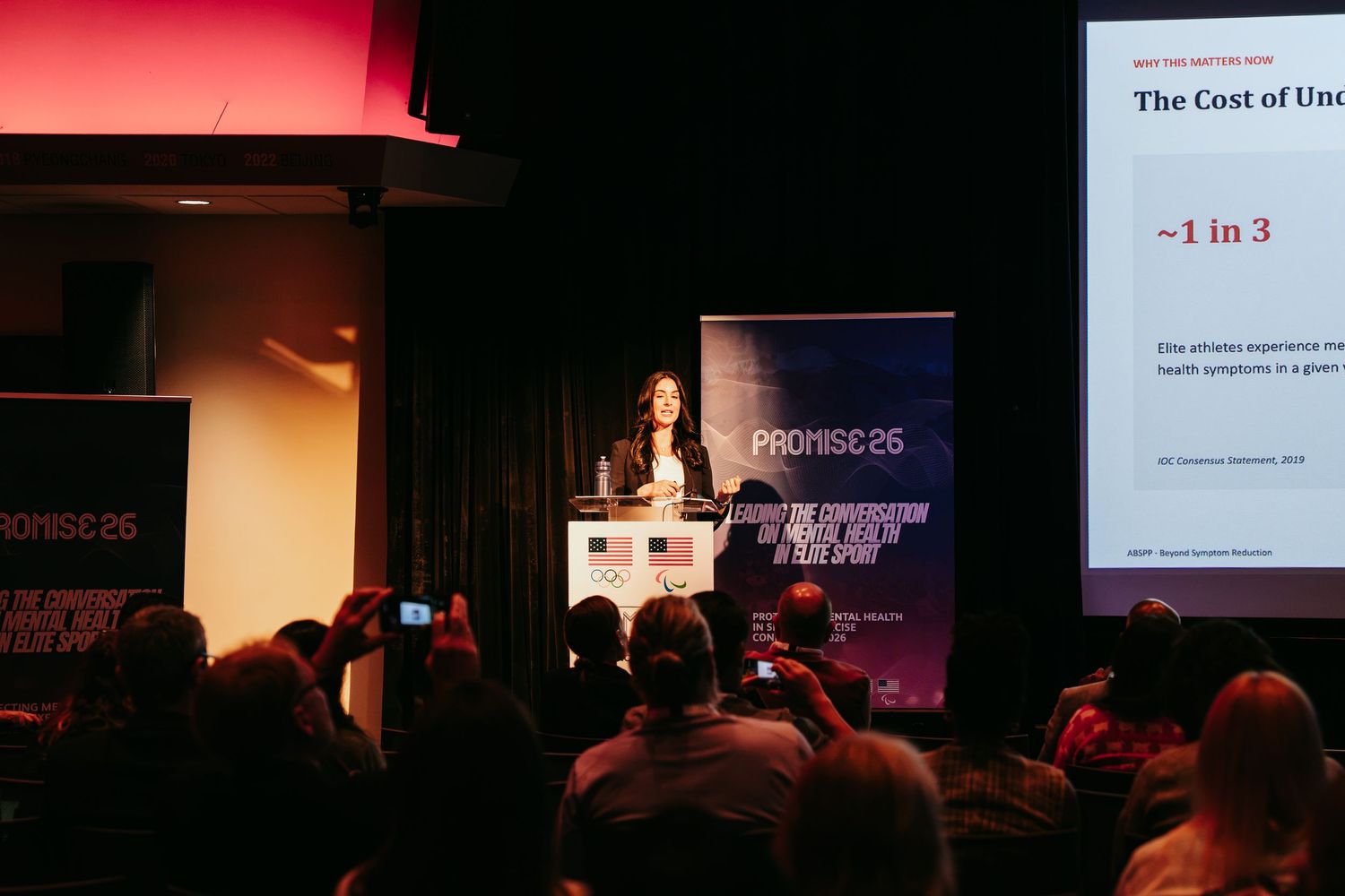 Speaker presents at a podium before an audience at a PROMISE 26 conference, with a slide on the cost of mental health issues.