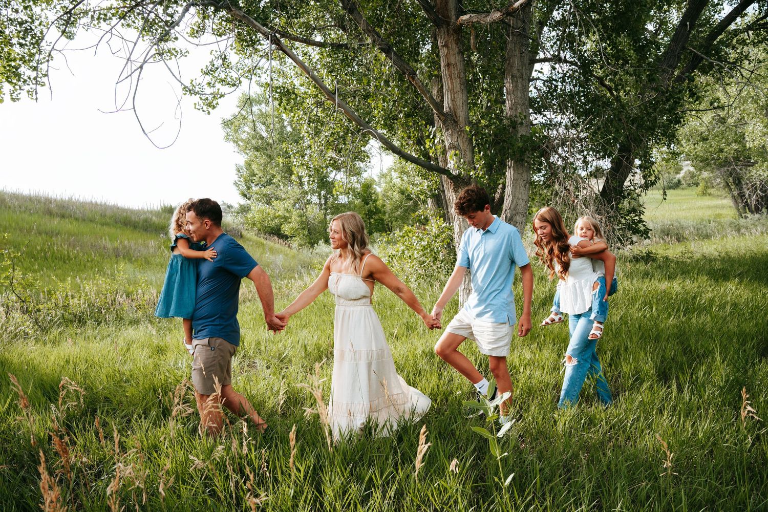 Family holding hands walking through a lush green meadow near trees on a sunny day.