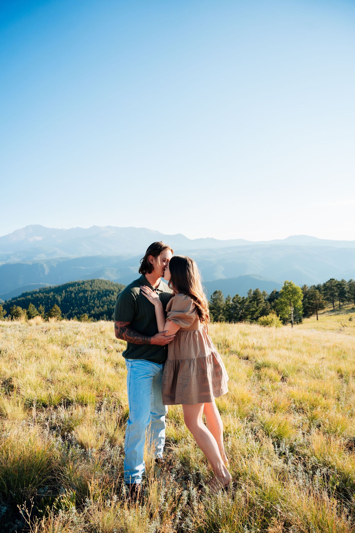 Couple embracing and kissing in a sunny alpine meadow with sweeping Rocky Mountain scenery in the background.