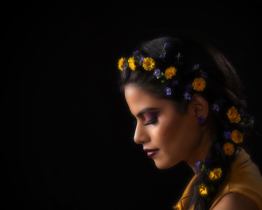 Side profile portrait against dark background with yellow flowers woven into dark hair in dramatic lighting.