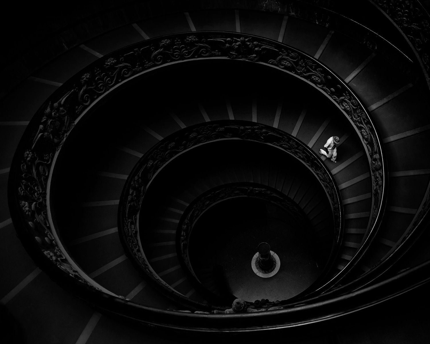 A dark spiral staircase photographed from above creates a mesmerizing circular pattern in black and white.