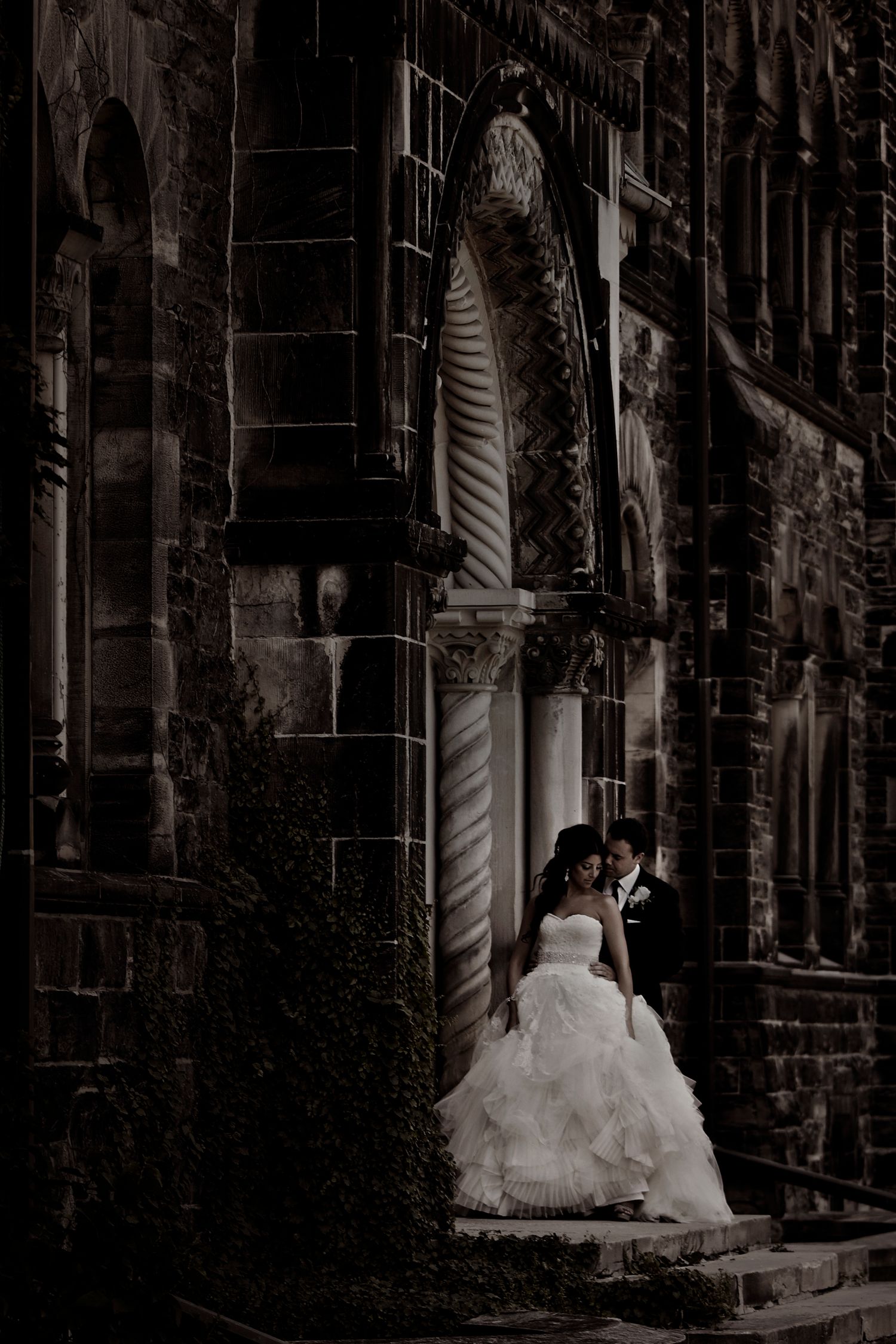 A couple in wedding attire stands against a dark stone building with dramatic shadows and lighting.