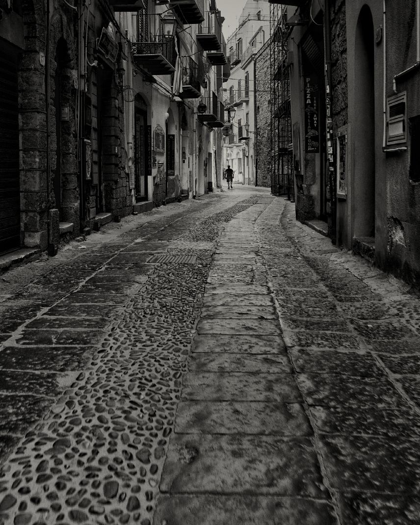 A narrow cobblestone street in an old Italian town with historic buildings and balconies in black and white.