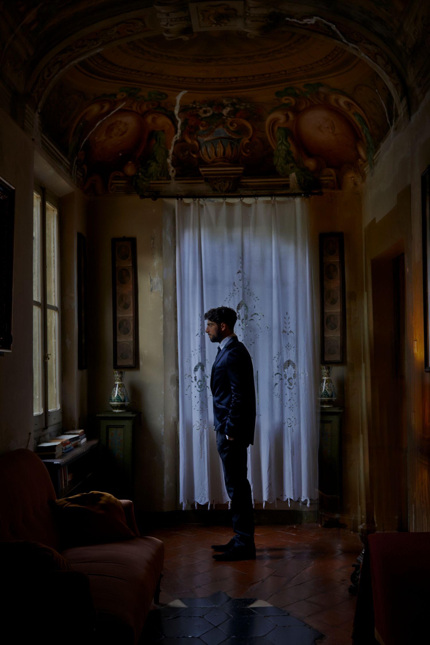 A silhouetted figure stands in a dark, abandoned room with sunlight filtering through sheer curtains on tall windows.