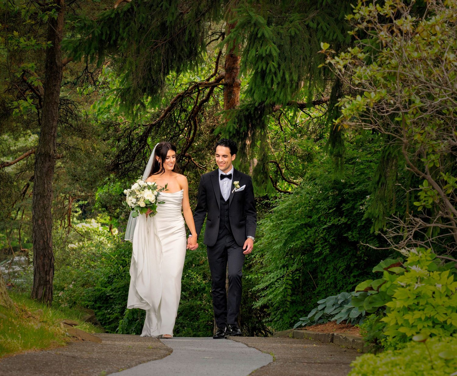 A couple in wedding attire walks together on a winding forest path surrounded by lush green trees.