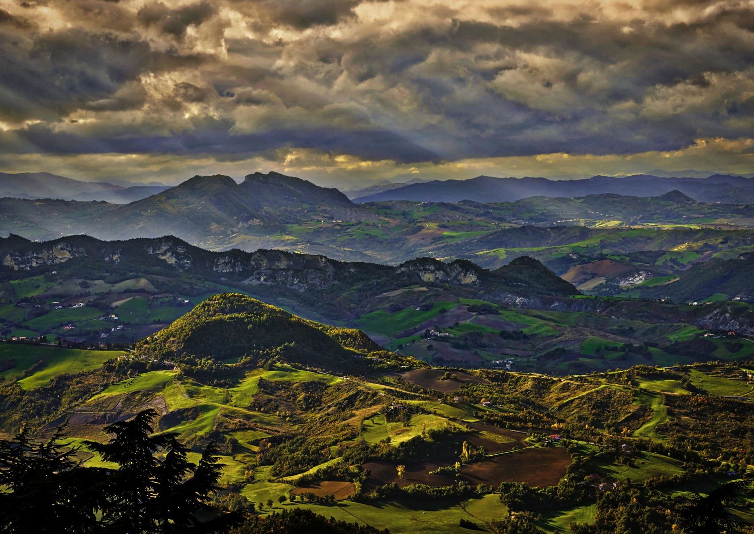 Dramatic panoramic view of green mountain ranges under stormy clouds at sunset with golden sunlight illuminating peaks.