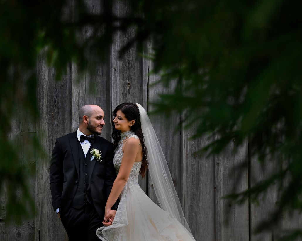 A couple in formal wedding attire share an intimate moment against a rustic wooden backdrop in a forest setting.
