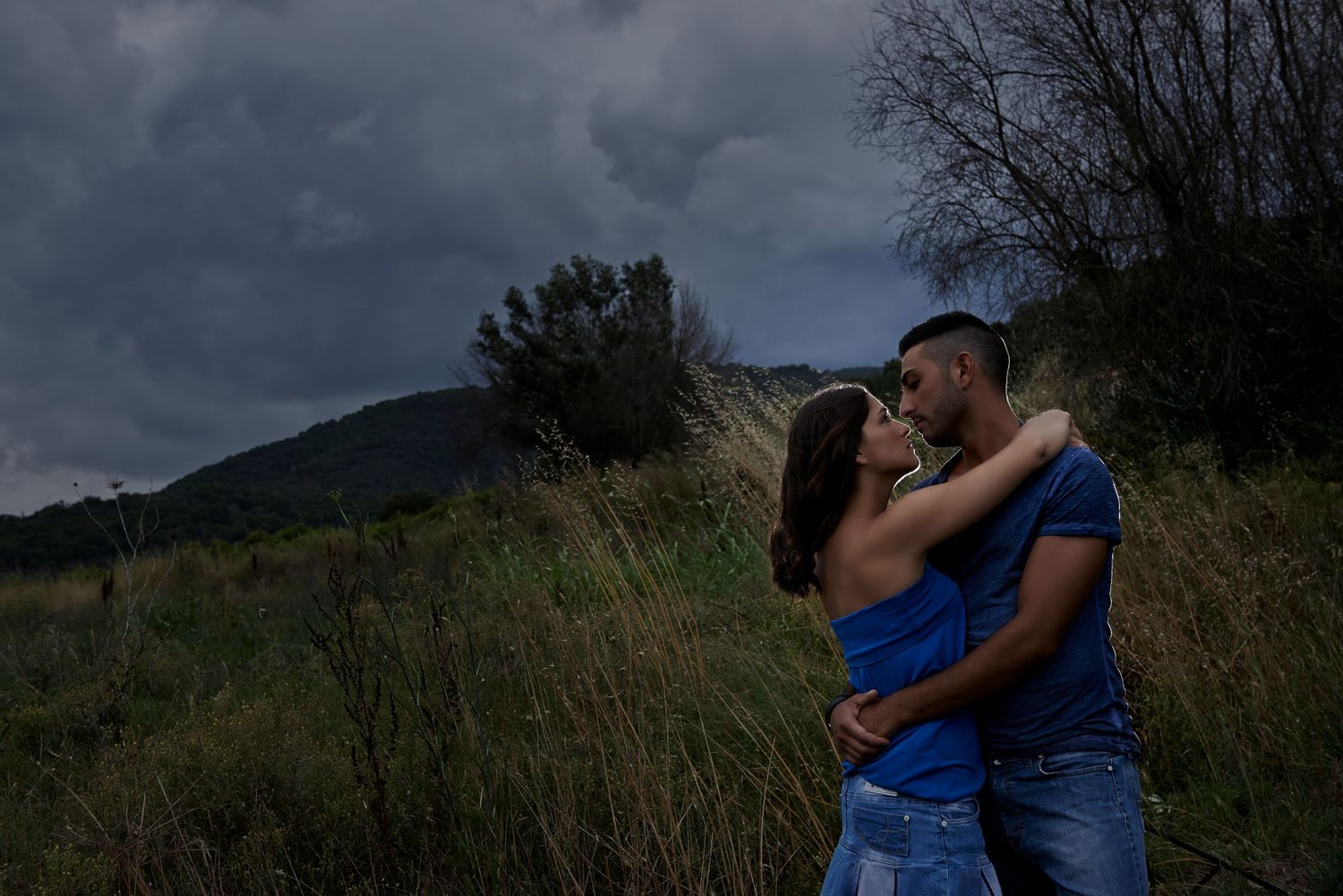 A couple in formal attire shares romantic moments in a grassy meadow with mountains in the background at sunset.
Horvath-Photography