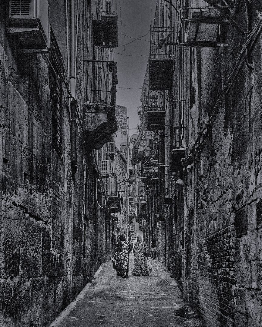 A black and white photograph of a narrow alleyway between tall buildings with laundry lines strung across it.