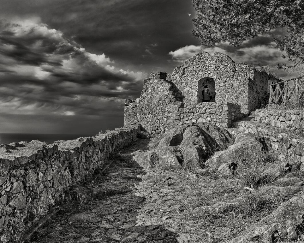 Black and white landscape of ancient stone ruins on a rocky hillside under dramatic cloudy skies.