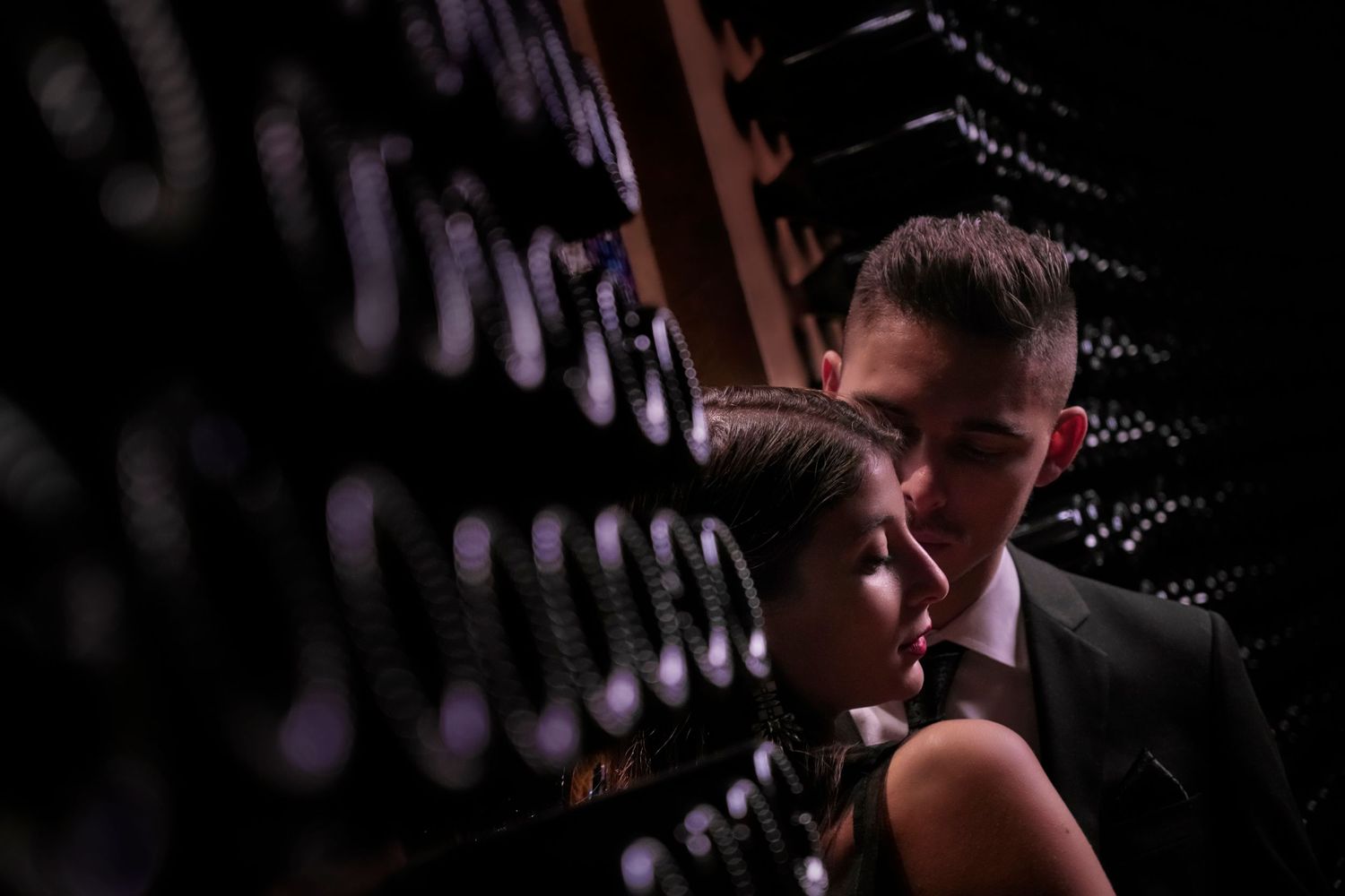 A romantic couple shares an intimate moment in a dimly lit wine cellar with bottles creating dramatic shadows.
Horvath-Photography