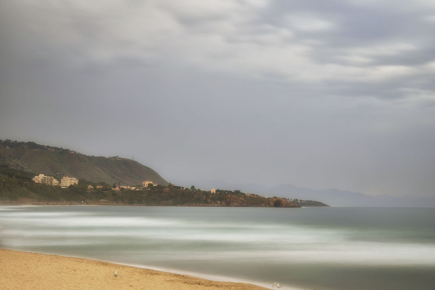 Panoramic view of a cloudy coastal landscape with rolling hills and a sandy beach stretching along the shoreline.