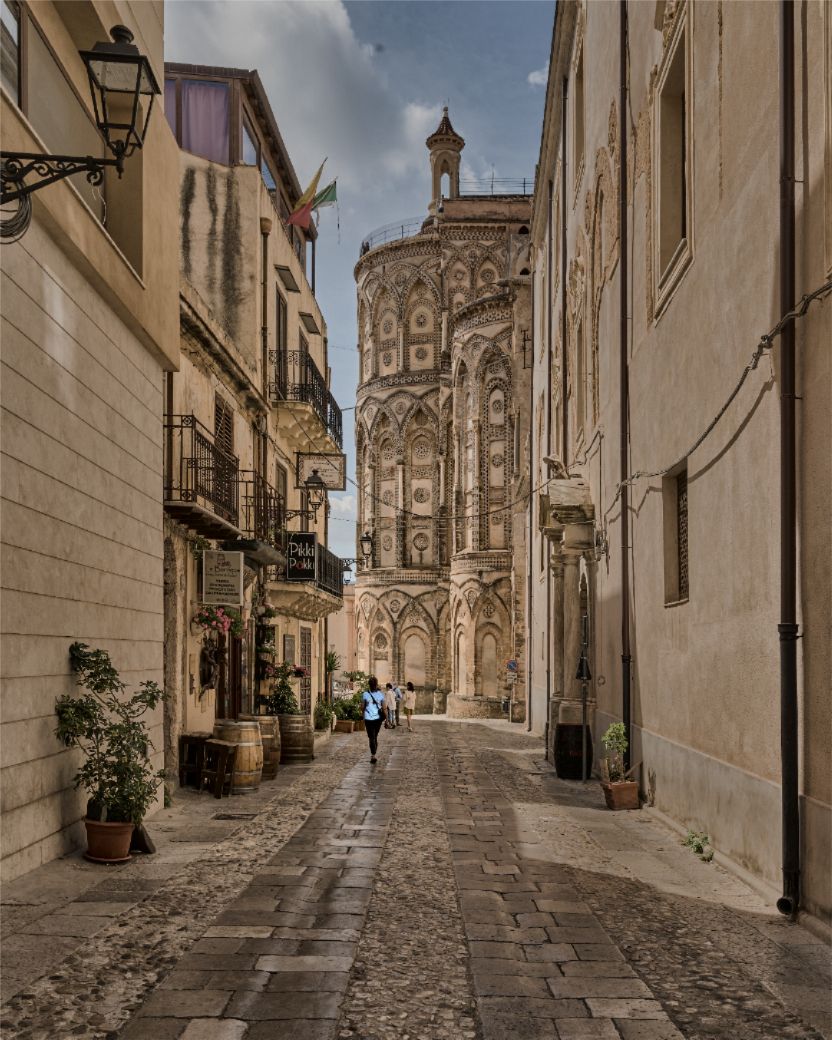 Narrow cobblestone street in an old European town leads to an ornate Baroque church facade with intricate stone carvings.