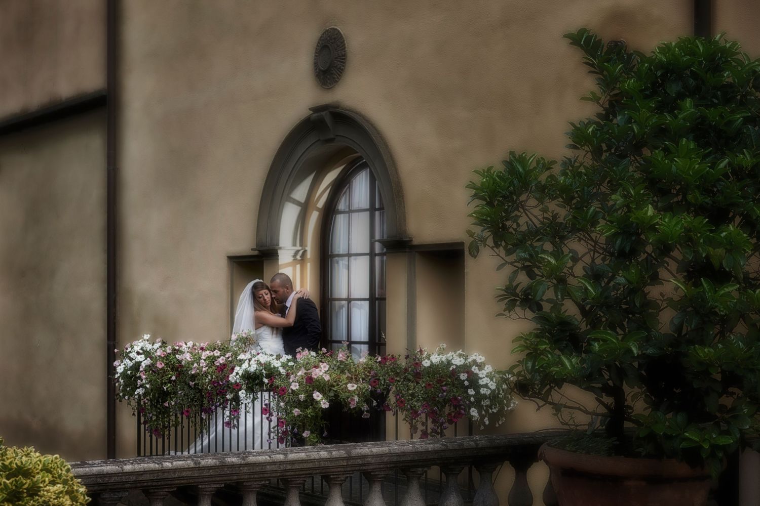 A romantic couple embraces on an elegant stone balcony with flowering planters and an arched doorway.