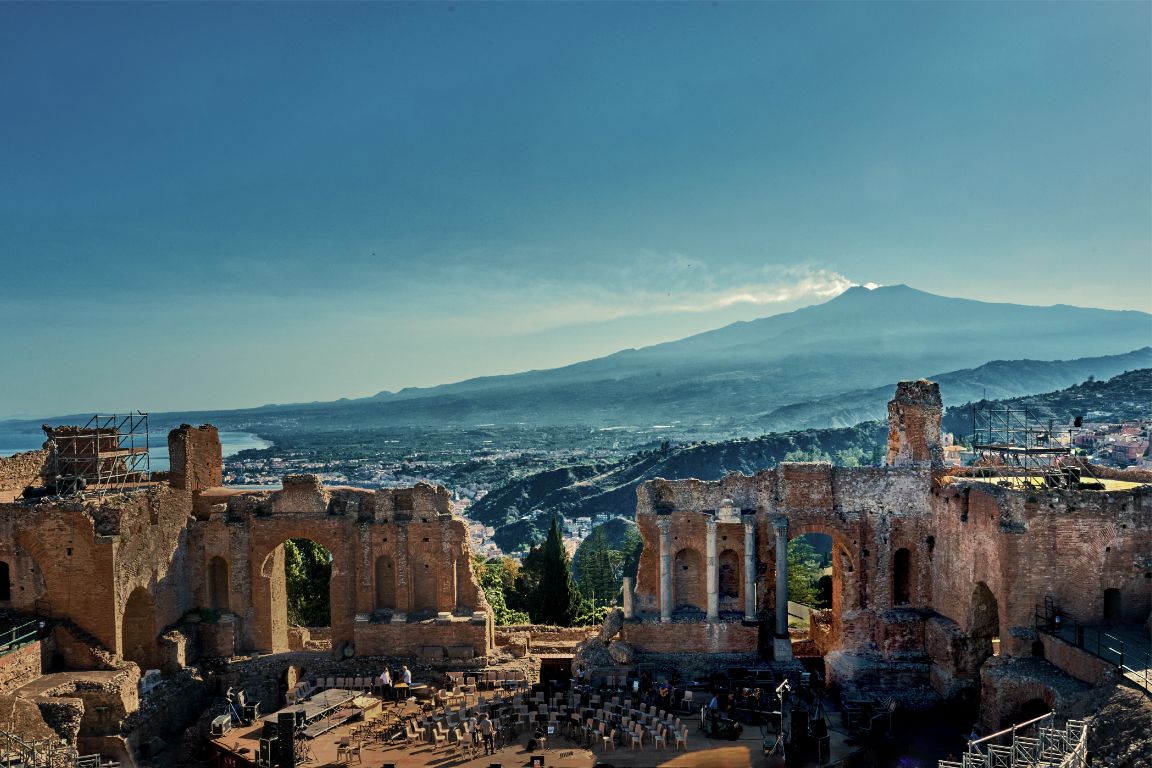 Panoramic view of Mount Etna and the historic city of Taormina in Sicily with ancient ruins in the foreground.